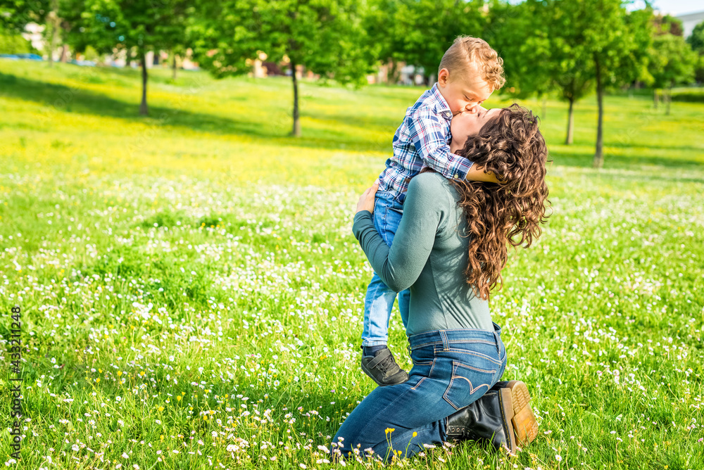 Fototapeta premium happy mom and child chilling in public park garden in summer. mother and son relaxing toghether outdoor sitting on grass playing. togetherness, love, family and happiness concept . sweet kiss portrait