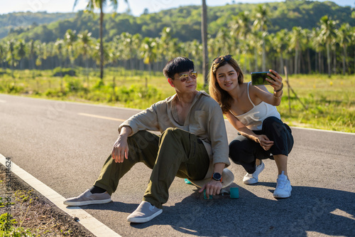 Young Asian Couple Lover using smartphone selfie themself while playing Surfskate on a Road, Summer Season