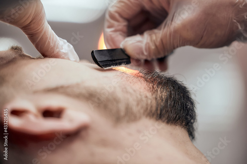 Professional barber shaves beard to young man using straight razor at the barbershop. Close-up, selective focus