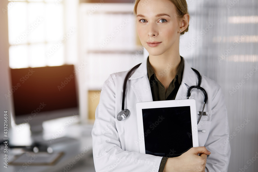 Cheerful smiling female doctor using tablet computer in sunny clinic. Portrait of friendly physician woman at work. Perfect medical service in hospital. Medicine concept Cheerful smiling female doctor using tablet computer in sunny clinic. Portrait of friendly physician woman at work. Perfect medical service in hospital. Medicine concept
