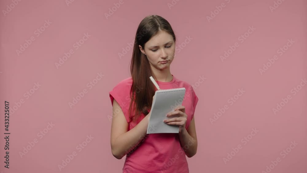 Portrait of fashion model is thinking and writing down her ideas in notebook. Young brown haired woman in pink t-shirt poses on pink studio background. Close up. Slow motion ready 59.94fps Portrait of fashion model is thinking and writing down her ideas in notebook. Young brown haired woman in pink t-shirt poses on pink studio background. Close up. Slow motion ready 59.94fps