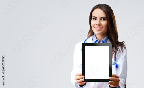 Portrait of happy smiling female doctor showing tablet pc with blank copy space area for slogan or text, over grey color background. Medical care concept. Selective focus.