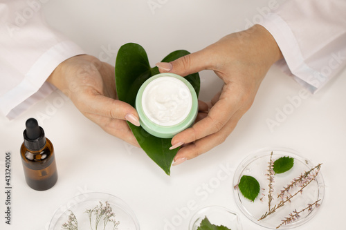 Close-up hands hold cosmetic jar with cream on fresh green leaf on white lab background. Dermatology, healthcare, treatment, microbiome skincare, plant-based probiotic products concept