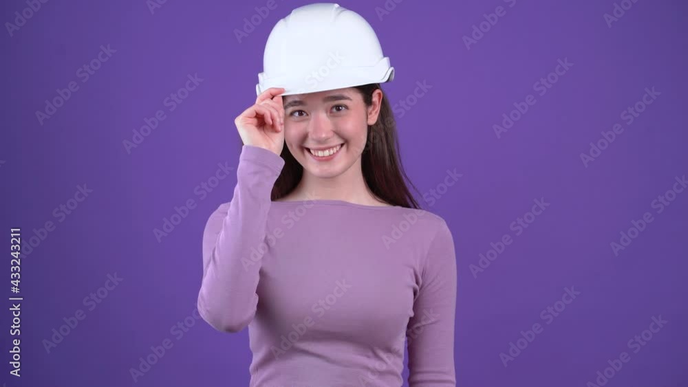 The mysterious young woman in the white helmet of the engineer looks up with her hand on her cap, then punches her with both fists, displays a wide smile. Brunette isolated on a purple background The mysterious young woman in the white helmet of the engineer looks up with her hand on her cap, then punches her with both fists, displays a wide smile. Brunette isolated on a purple background