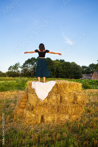 Young beautiful woman stands on a large pile of straw bales. Freedom, wind, setting sun.