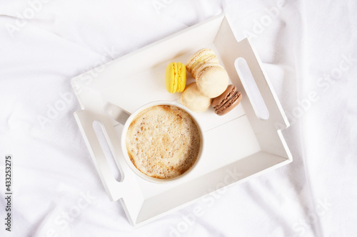 Cup of coffee, macaroons on the square tray in the middle on the white bedsheet. Service. Holiday. Morning surprise. Coffee break atmosphere. 