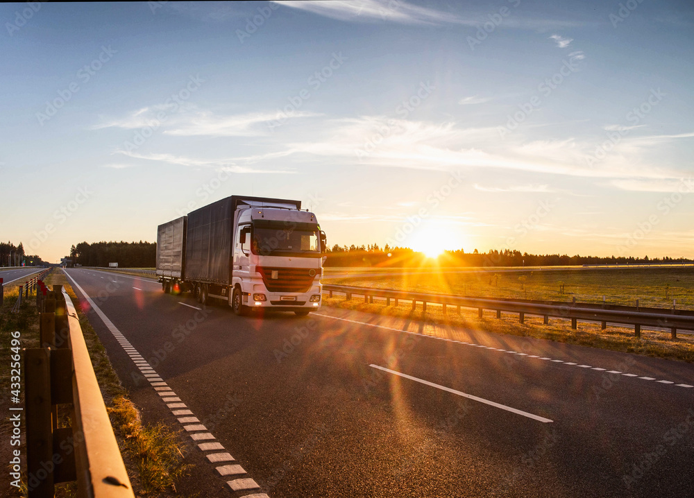 A white truck with a semitrailer carries cargo along the highway against the backdrop of a sunny sunset in the evening. Business and freight concept, copy space for text A white truck with a semitrailer carries cargo along the highway against the backdrop of a sunny sunset in the evening. Business and freight concept, copy space for text