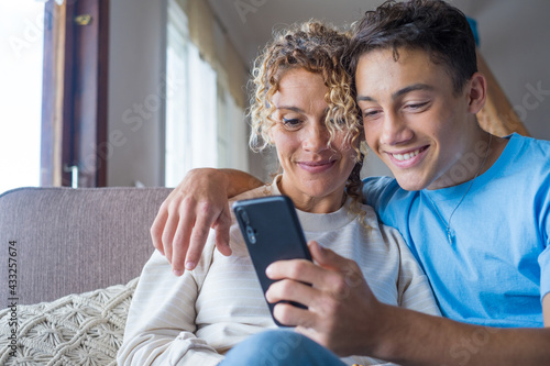 Smiling middle-aged 40s mother rest with grown-up son using smartphone together, happy young man enjoy family weekend with mom browsing wireless Internet on cellphone, have fun at home
