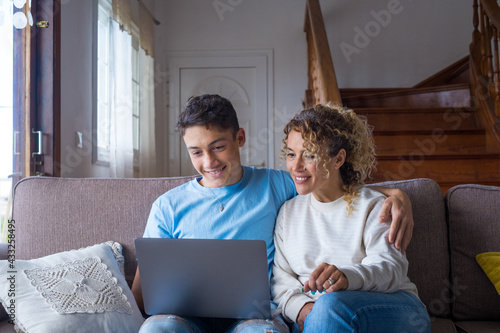 Adult son and middle-aged 40s mother sitting on couch in cozy living room holding on lap notebook laughing on funny comedy movie  enjoy on-line amusements websites  internet virtual fun online concept