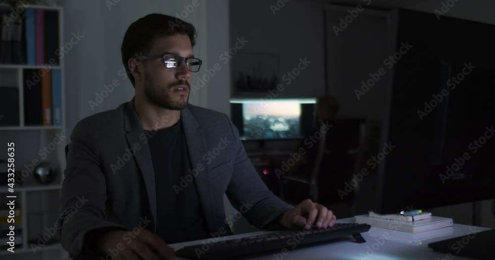 Lateral dolly of young caucasian handsome businessman in eye glasses late at night at office in front of computer. Male broker following rates and bets. Focused man working late. Lateral dolly of young caucasian handsome businessman in eye glasses late at night at office in front of computer. Male broker following rates and bets. Focused man working late.