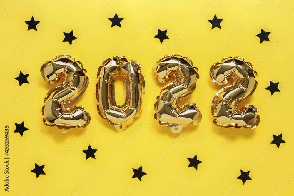 Fototapeta premium Christmas or New Year background. Text 2022, made of golden balloons, on a yellow background, with black confetti stars. Postcard or calendar. Merry Christmas or Happy New Year.