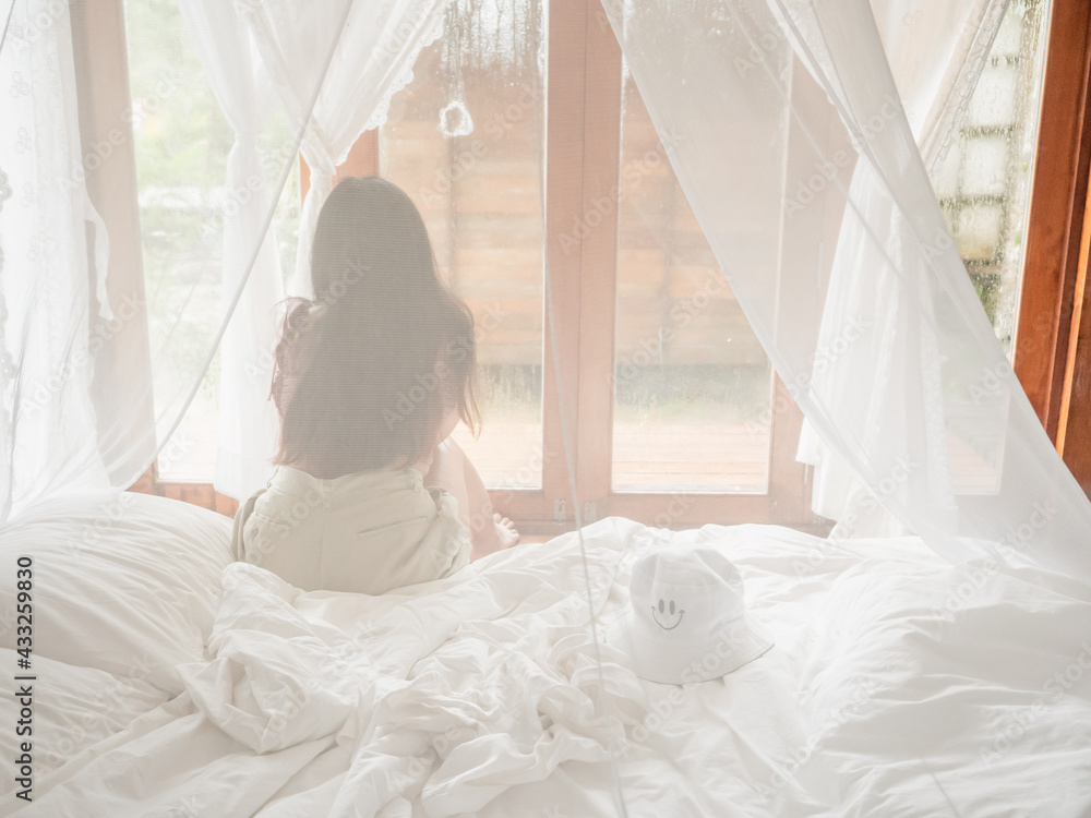 Back view of woman relaxing sitting on bed in mosquito net of an hotel room looking through window in vacations Back view of woman relaxing sitting on bed in mosquito net of an hotel room looking through window in vacations