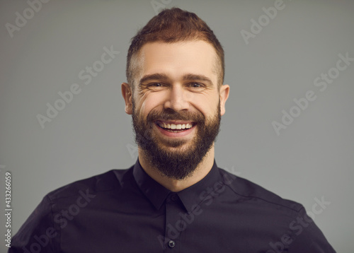 Headshot of cheerful businessman in black dress shirt with positive beaming smile on friendly open face. Studio portrait of happy stylish bearded young man with modern short haircut with shaved sides