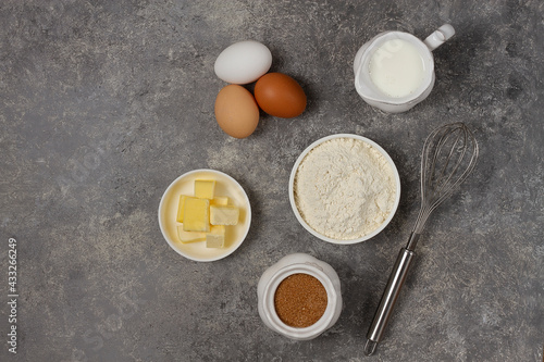 Dough ingredients, milk, eggs, flour and whisk, butter, brown cane sugar, on a gray table, top view, background, no people, horizontal. 
