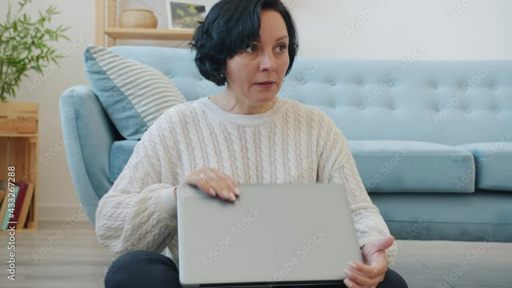 Smiling mature woman is using laptop typing then turning off computer and thinking sitting on floor indoors at home. Modern technology and lifestyle concept. Smiling mature woman is using laptop typing then turning off computer and thinking sitting on floor indoors at home. Modern technology and lifestyle concept.