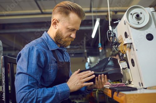 Male worker checking quality of footwear. Side view of man sitting at table with shoe sewing machine, holding black men's leather boot and looking at seams and stitches. Manufacturing industry concept