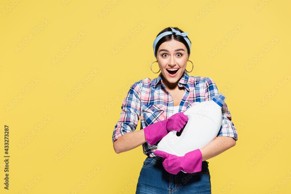 Surprised woman in rubber gloves holding bottle of detergent isolated on yellow Surprised woman in rubber gloves holding bottle of detergent isolated on yellow