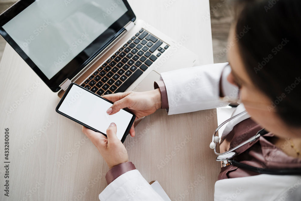 © Eva Blanco/Westend61 - Female doctor using mobile phone by laptop at desk © Eva Blanco/Westend61 - Female doctor using mobile phone by laptop at desk