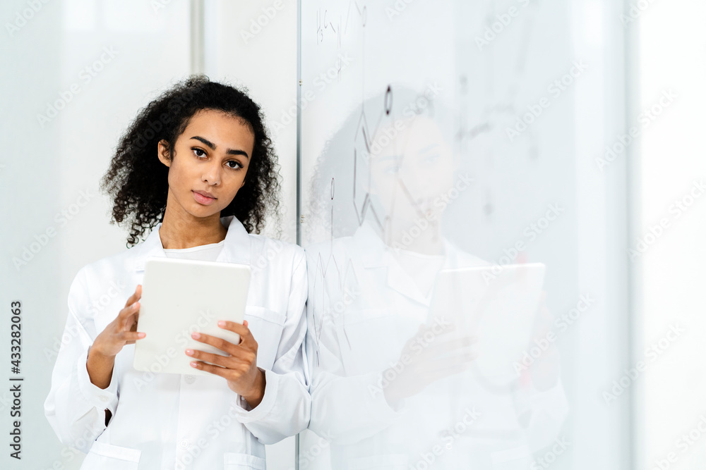 Confident female researcher holding digital tablet while leaning on whiteboard in laboratory Confident female researcher holding digital tablet while leaning on whiteboard in laboratory