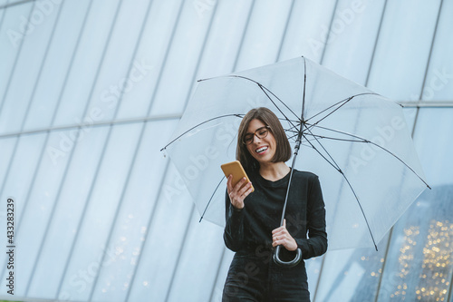 Cheerful young woman with umbrella using smart phone while standing in front of glass building photo