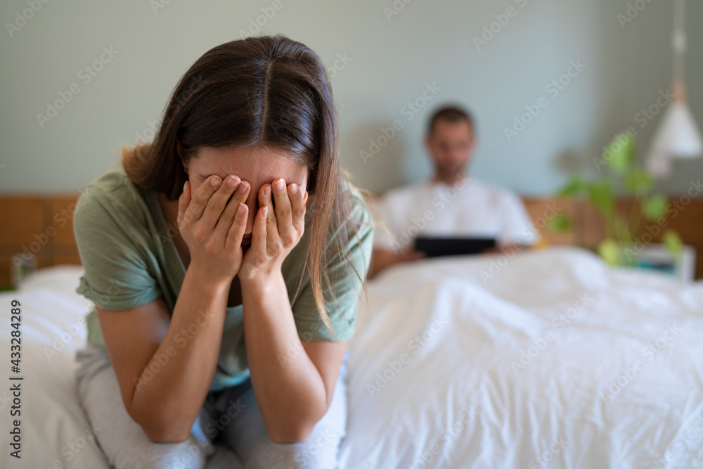Desperate woman covering her face sitting on bed while boyfriend using digital tablet in background at home Desperate woman covering her face sitting on bed while boyfriend using digital tablet in background at home