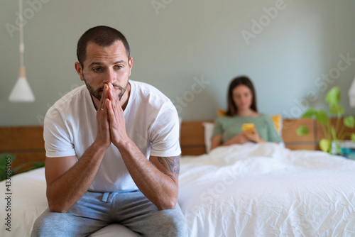 Worried young man sitting on bed while girlfriend using smart phone in background at home