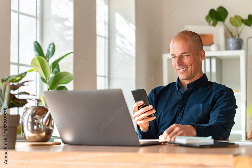 Smiling businessman with laptop using mobile phone while working at home Smiling businessman with laptop using mobile phone while working at home