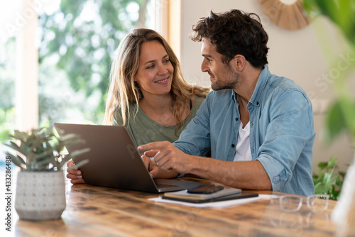 Smiling couple looking at each other while sitting in front of laptop at table