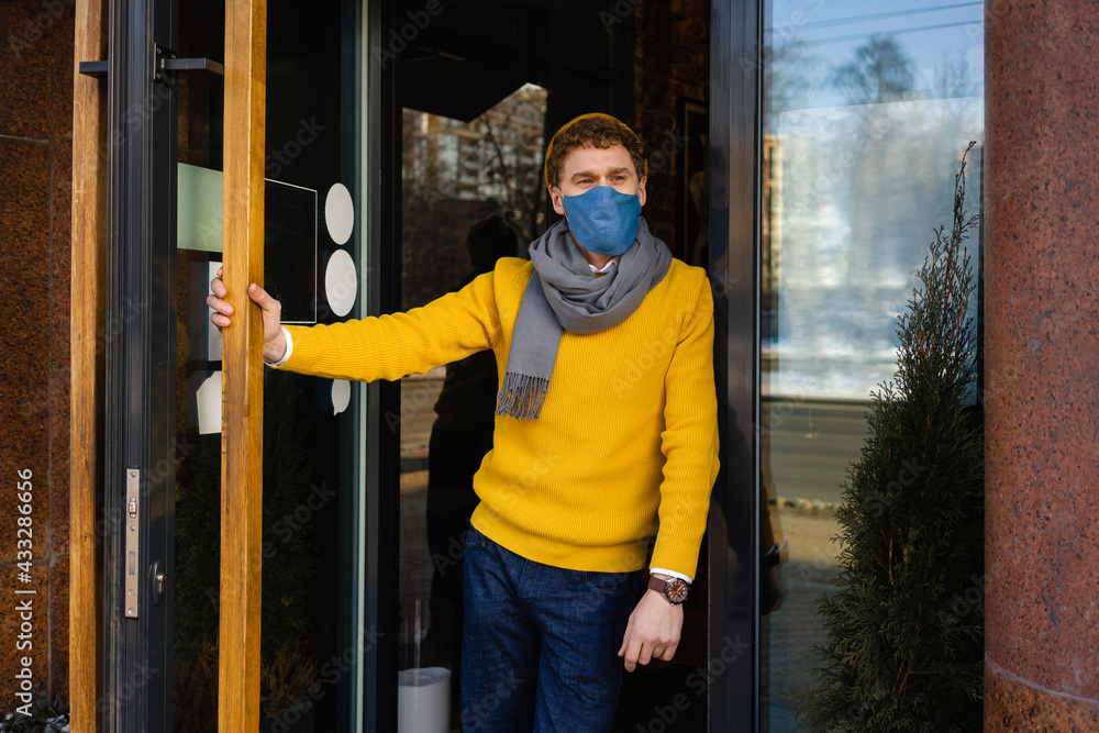Male owner wearing protective face mask leaning at cafe entrance during COVID-19 Male owner wearing protective face mask leaning at cafe entrance during COVID-19