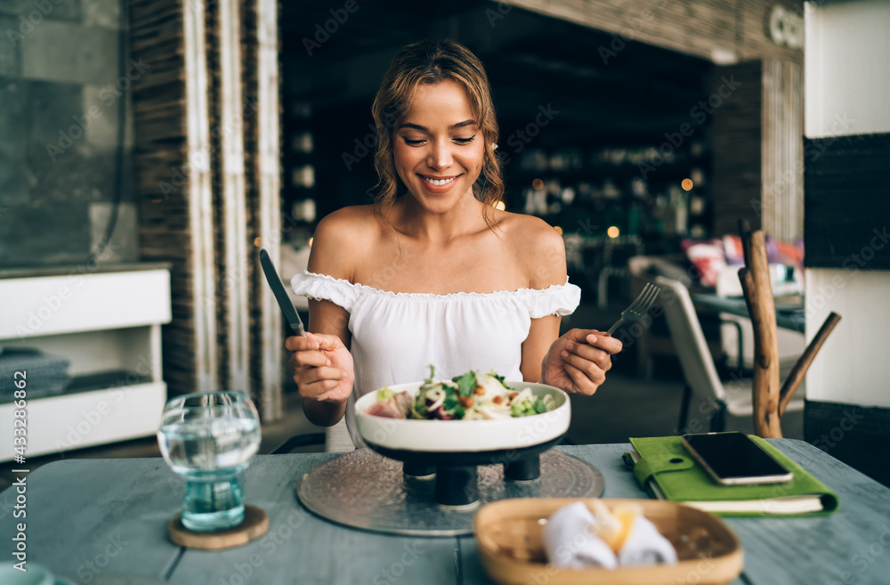 Positive woman with cutleries eating salad in modern restaurant