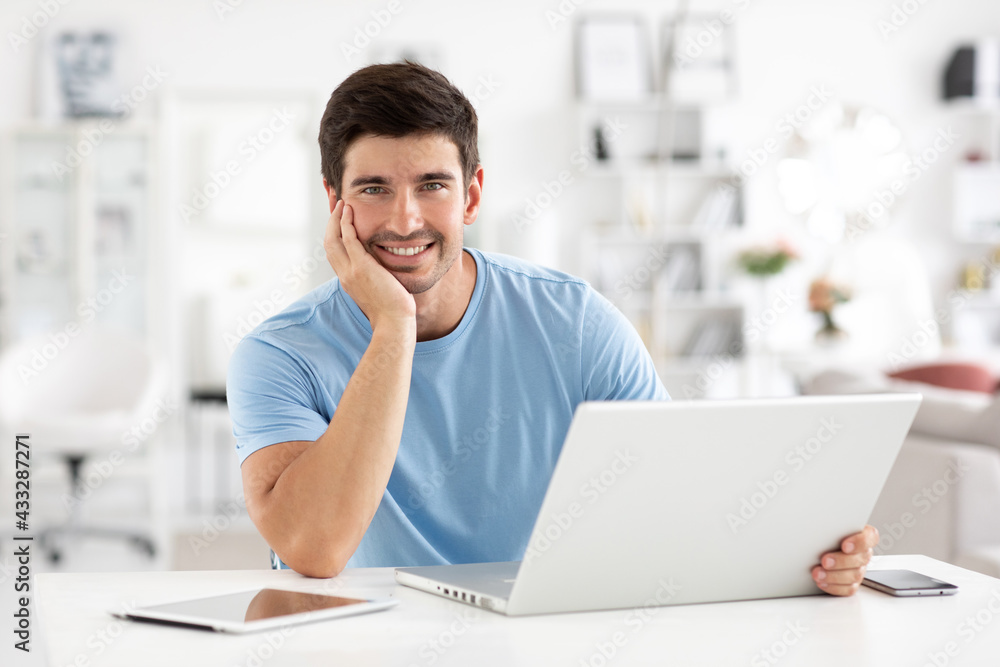 Handsome cheerful young male copywriter uses modern laptop for work, wears a blue t-shirt, look at camera Handsome cheerful young male copywriter uses modern laptop for work, wears a blue t-shirt, look at camera