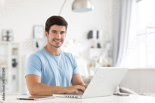 Handsome cheerful young male copywriter types information for advertisment of website, uses modern laptop for work