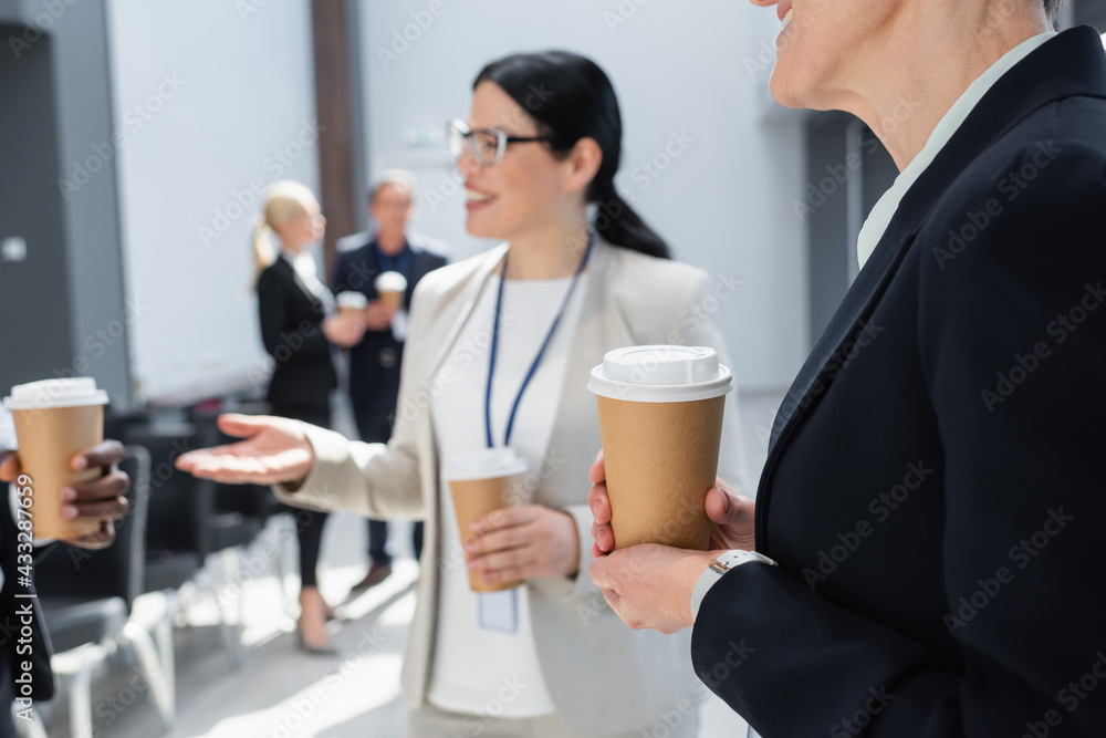 businesswoman holding coffee to go near colleagues talking on blurred background