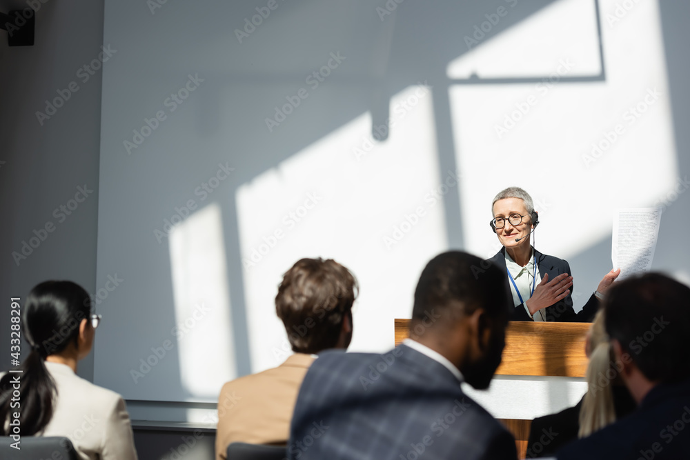 smiling lecturer pointing at document near blurred business people during seminar
