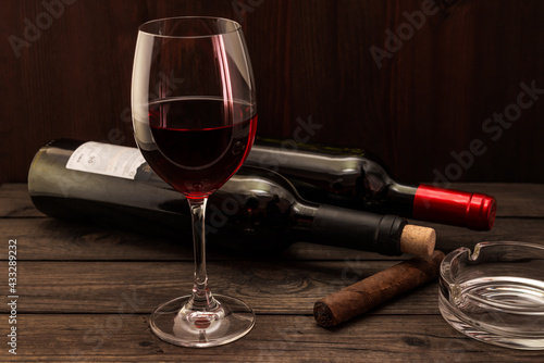 Two bottles of red wine with a glass and cuban cigar with ashtray on an old wooden table. Focus on the cuban cigar
