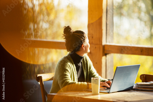 Woman working at laptop looking out window in sunny cafe