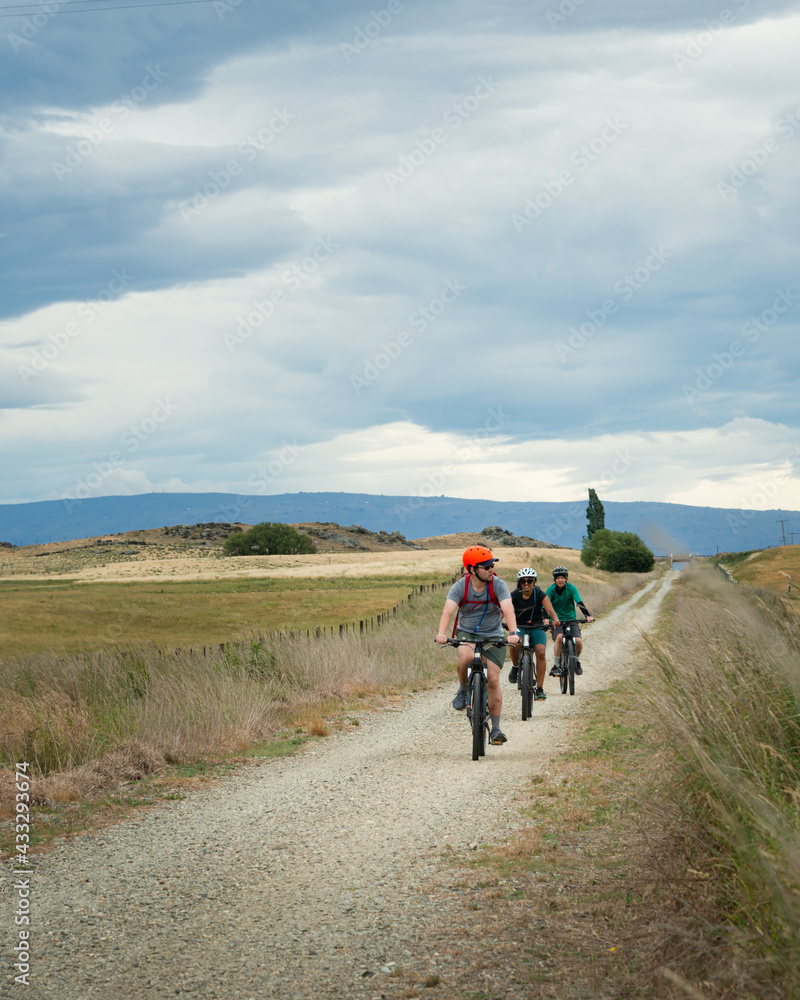 Three cyclists riding the Otago Central Rail Trail under the fluffy white clouds, South Island. Vertical format.