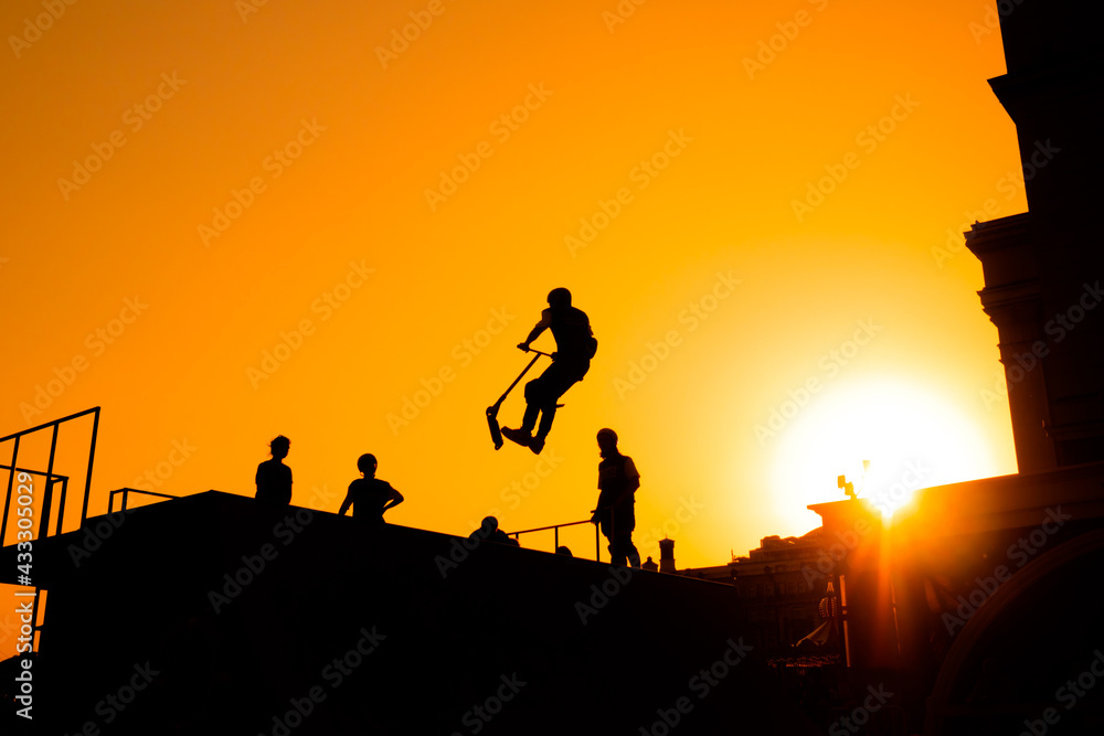Unrecognizable teenage boy silhouette showing high jump tricks on scooter against orange sunset sky at skatepark. Sport, freestyle, extreme, youth, urban culture, outdoor activity concept Unrecognizable teenage boy silhouette showing high jump tricks on scooter against orange sunset sky at skatepark. Sport, freestyle, extreme, youth, urban culture, outdoor activity concept