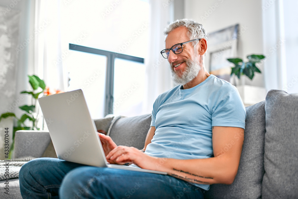A mature man is sitting on the couch with a laptop on his lap at home. Freelance, work online. The man is texting on his laptop. A mature man is sitting on the couch with a laptop on his lap at home. Freelance, work online. The man is texting on his laptop.