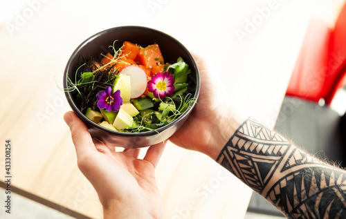 A man holds a bowl of Poke salad with salmon, avocado, cucumber, rice, Tamarind sauce, mango sauce, nori, lime, sesame, cilantro. Asian seafood salad concept.