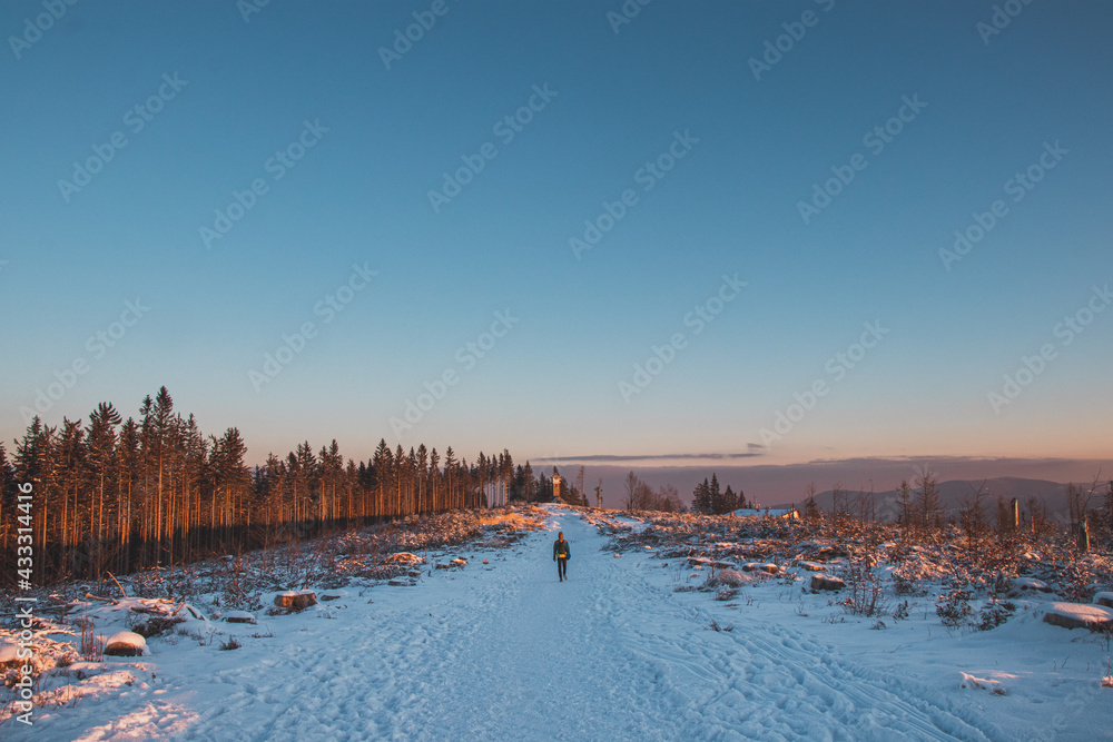 Panorama of a beautiful winter landscape on top of a hill and a well-trodden path and a traveler passing it along this path at sunrise. Aerial blue sky with orange tone illuminates white snow