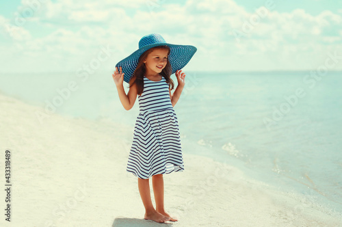 Summer portrait beautiful little girl child wearing a striped dress and straw hat on a beach on a sea background