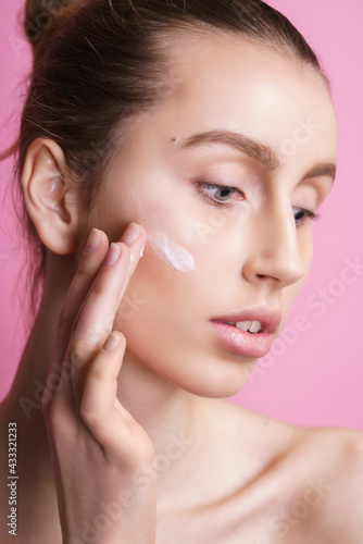 Beautiful young woman applying cream on her face against color background