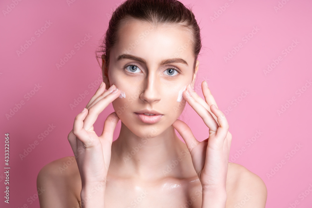 Beautiful young woman applying cream on her face against color background Beautiful young woman applying cream on her face against color background