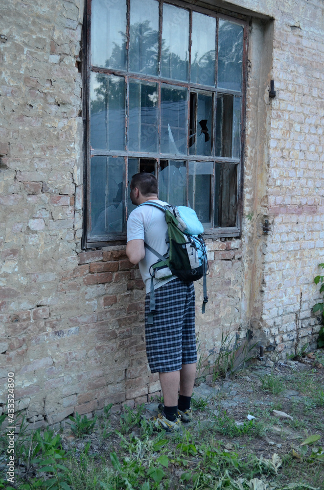 A man with a backpack on his back looks through a broken window into an abandoned house