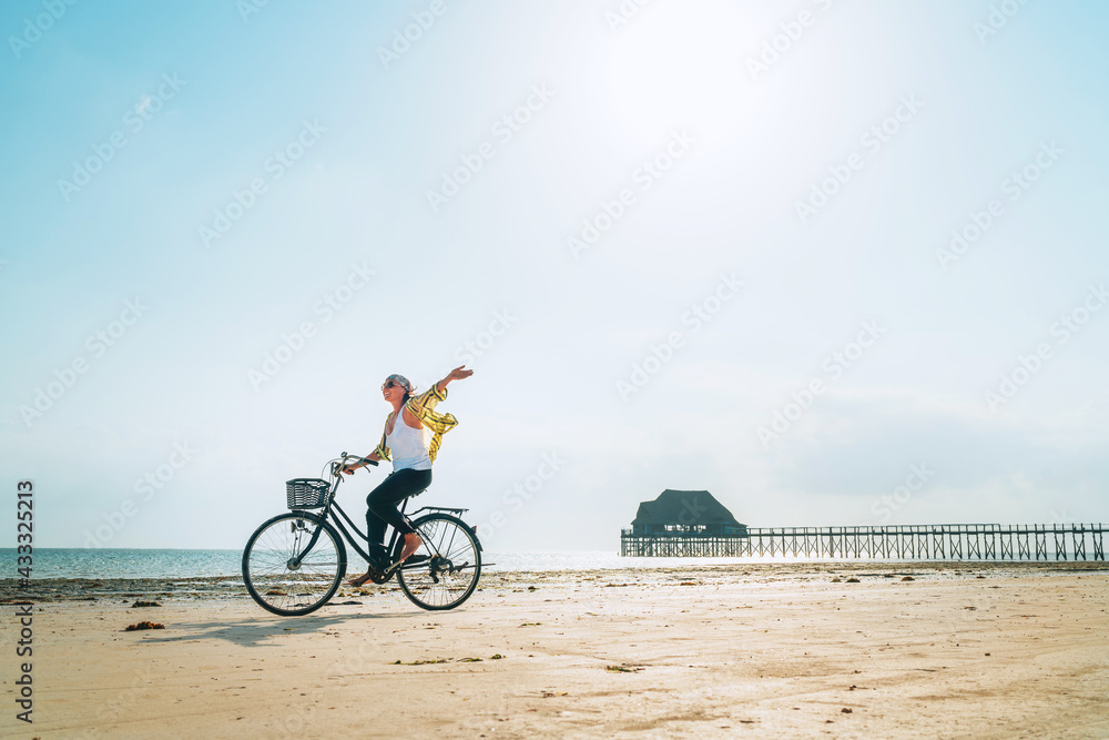 Fototapeta premium Kiwengwa beach on Zanzibar island, Tanzania.Young female dressed light summer clothes joyfully threw up her hand riding old vintage bicycle with front basket on the low tide ocean white sand coast on