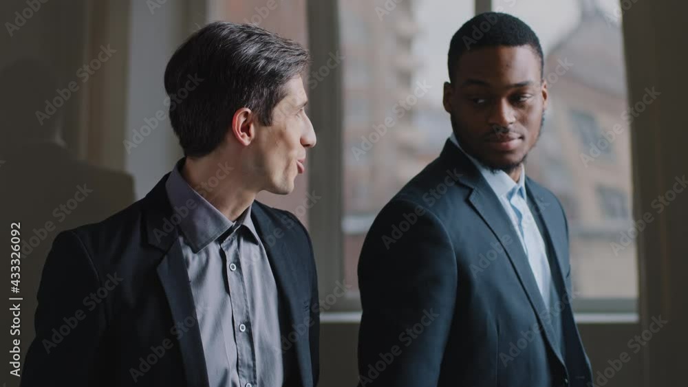 Two coworker colleagues stand in modern company office look out window, happy caucasian business man talks points direction with forefinger outside boss african ethnic black leader manager nods yes Two coworker colleagues stand in modern company office look out window, happy caucasian business man talks points direction with forefinger outside boss african ethnic black leader manager nods yes