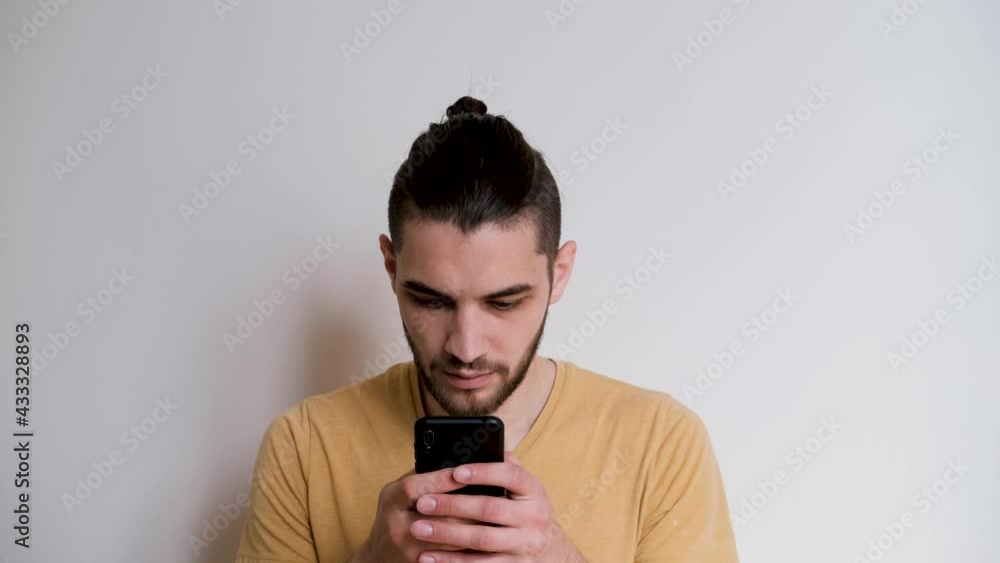 Young handsome man of Caucasian European appearance with beard and ponytail chats with mobile phone and looks carefully. Online correspondence using smartphone from home against white wall. Young handsome man of Caucasian European appearance with beard and ponytail chats with mobile phone and looks carefully. Online correspondence using smartphone from home against white wall.