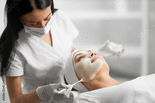 Side view of beautiful brunette beautician applying special white cream for face young cute patient. Concept of process procedure for improving face skin with cosmetics. photo
