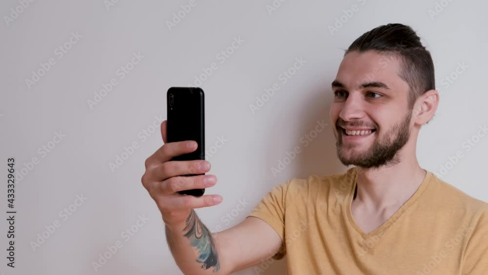 Online conference from home on white wall background. Young handsome man of Caucasian European appearance with beard and ponytail is talking on video link using mobile phone and is emotionally happy. Online conference from home on white wall background. Young handsome man of Caucasian European appearance with beard and ponytail is talking on video link using mobile phone and is emotionally happy.